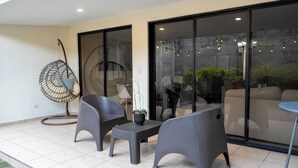 A patio area with wicker furniture, a hanging chair, and a view through a glass door to an outdoor space with plants and a sofa. - Family Oasis w/ Full A/C • Pools & 24/7 Security (Apopa)