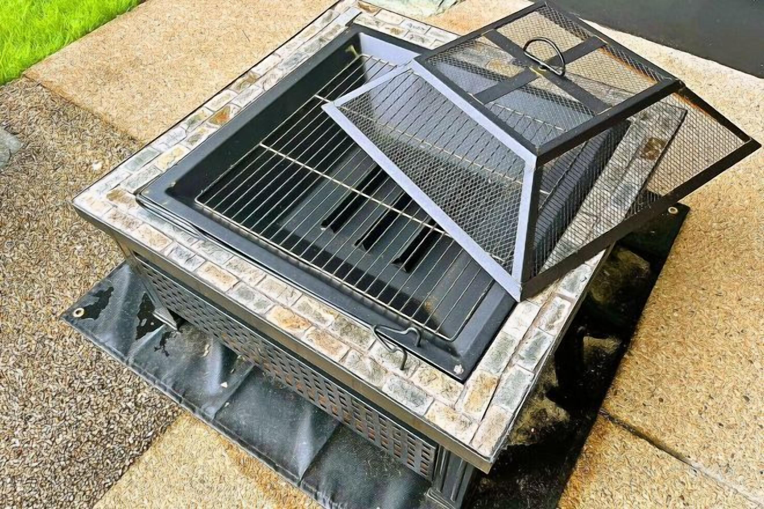A barbecue grill with a metal grate and a mesh cover, placed on a stone base.