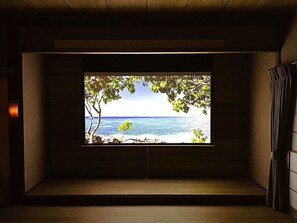 Quarto com uma janela grande de frente para uma praia, uma mesinha e uma cortina à direita.