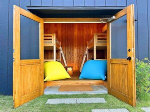 Une structure en bois avec une porte ouverte, laissant entrevoir une pièce dont l’intérieur est également en bois et qui contient deux coussins aux couleurs vives.