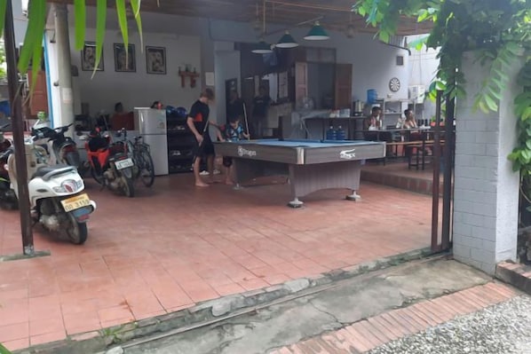 Una mesa de billar en una habitación con piso de baldosas, un refrigerador y una motocicleta estacionada afuera.