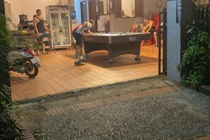 Une table de billard dans une pièce dont le sol est carrelé, une moto garée à l’extérieur, et une personne qui joue.