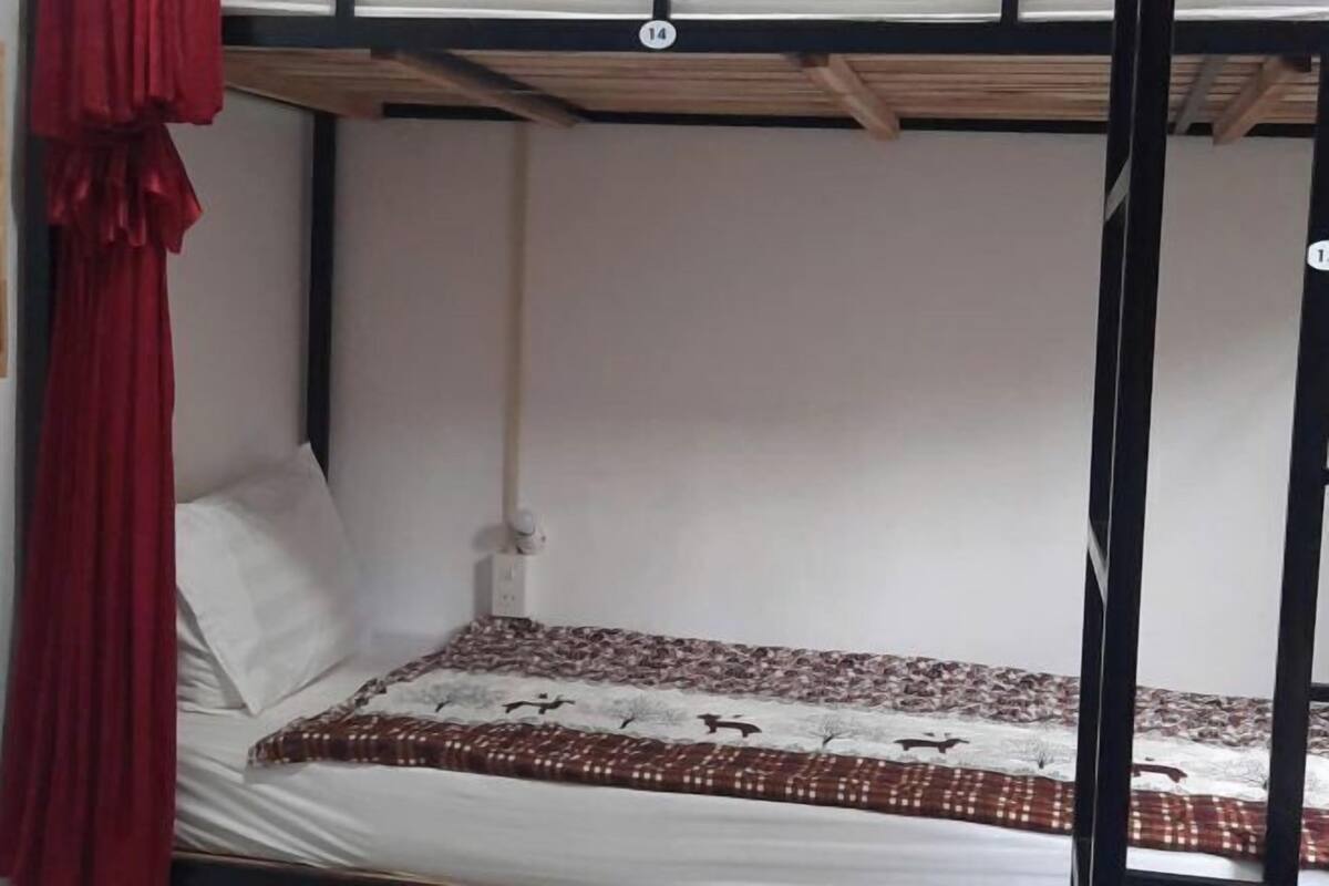 A single bunk bed with a patterned bedspread, a white pillow, and a red curtain.