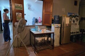 A kitchen with a refrigerator, a fan, and a table with cups and a kettle. - Aurora Backpackers Hostel (Luang Prabang)