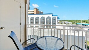 Un balcon avec une table et des chaises en verre, donnant sur une rue bordée d’immeubles et offrant une vue sur un ciel dégagé.