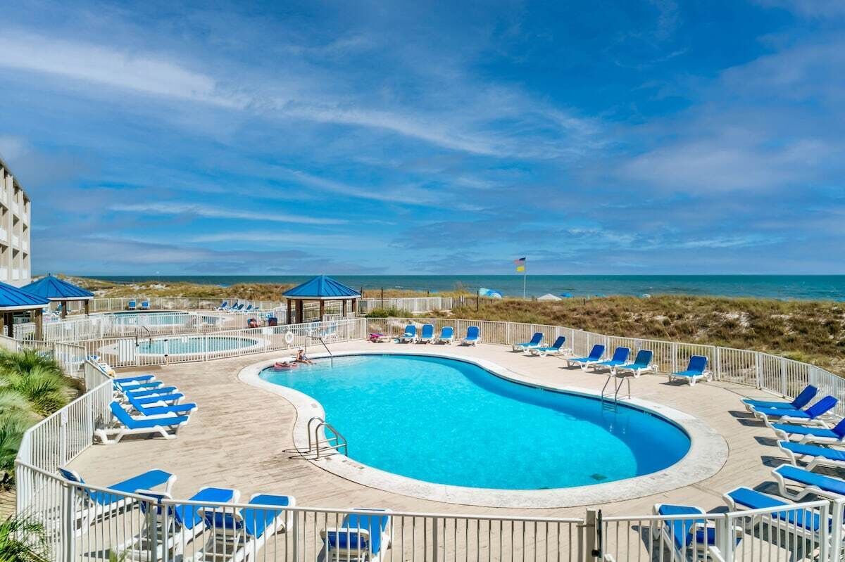 Zona de piscina con sillones reclinables azules y un pabellón, rodeada por una valla, con vistas a la playa.