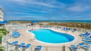 Un espace piscine avec des chaises longues bleues et un kiosque, entouré d’une clôture, offrant une vue sur la plage.
