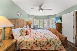 Un dormitorio con cama, ventilador de techo, televisión, cómoda y ventana con persianas.