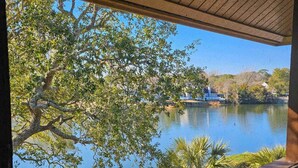 A view from a covered area overlooking a calm lake with a boat docked on the right, surrounded by trees and a distant building. - Laurel Ct 307 in Kingston w/Elevator, Pool, Beach (Myrtle Beach)