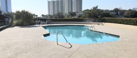 A swimming pool with a metal ladder, surrounded by a concrete deck and a hedge. In the background, there are tall residential buildings.