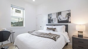 A modern bedroom with a large bed, a desk, and a chair. There are two abstract paintings on the wall and a window with a view of buildings.