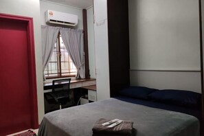 A bedroom with a bed, a desk with a chair, a window with curtains, and an air conditioning unit. - Putrajaya Transit Romestay (Putrajaya)