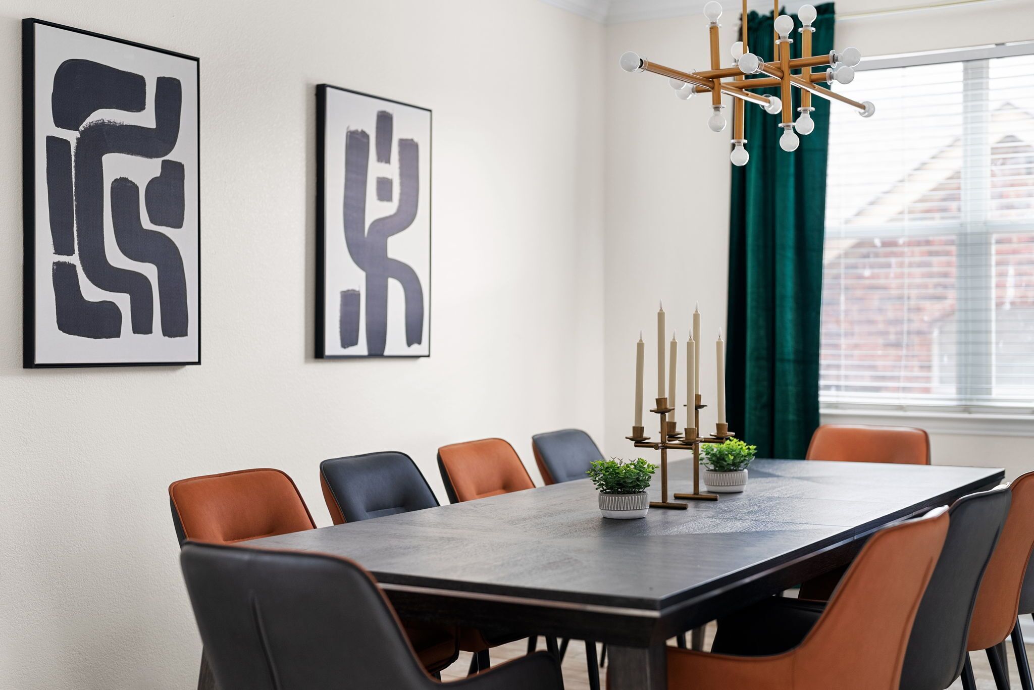 Een eethoek met een rechthoekige tafel, zes stoelen, twee abstracte schilderijen, een potplant, een kandelaar en een raam met gordijnen.