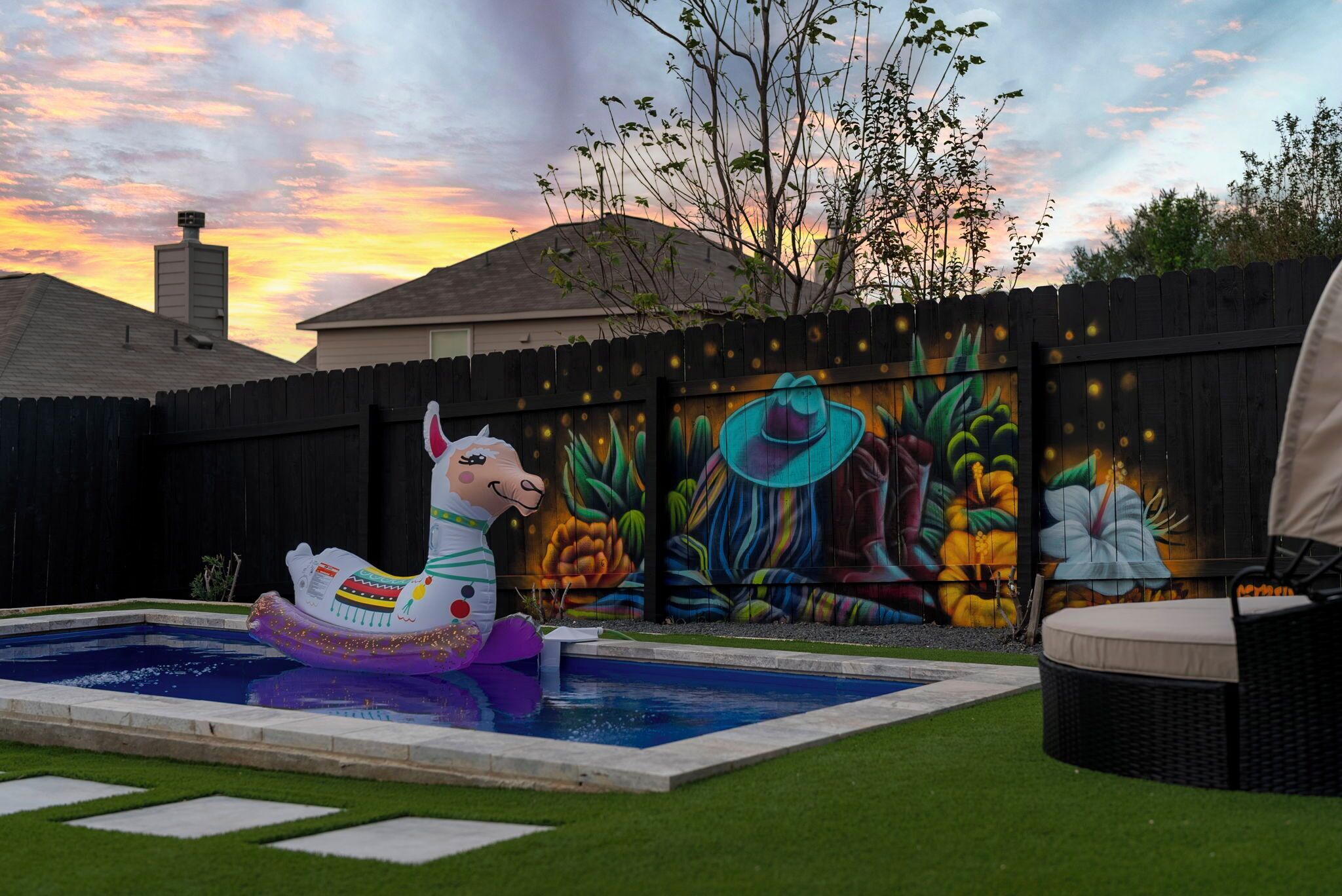 A backyard with a swimming pool, a colorful inflatable llama, and a mural on the fence.
