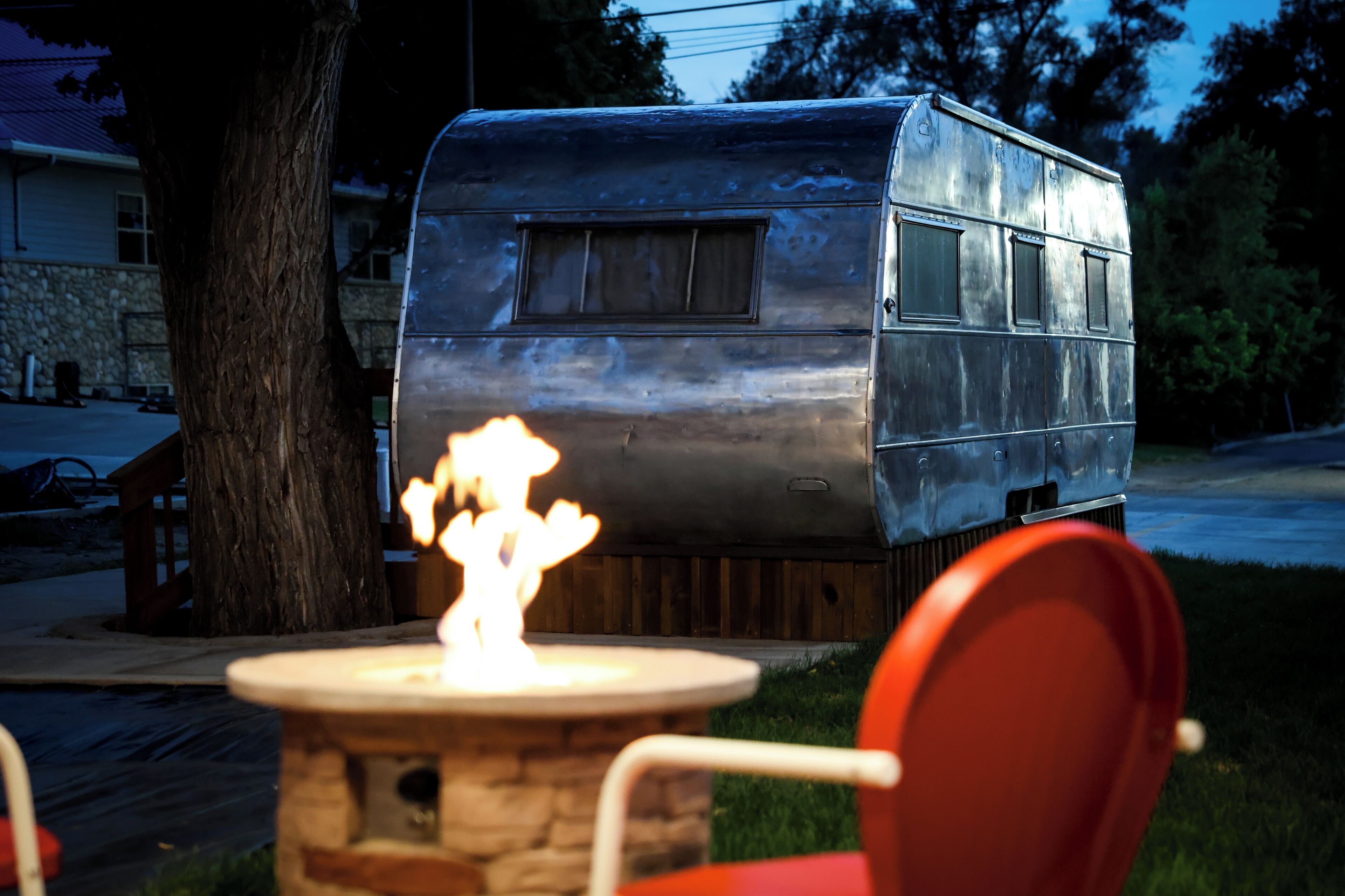 A vintage trailer with a fire in front of it, a tree, and a house in the background.