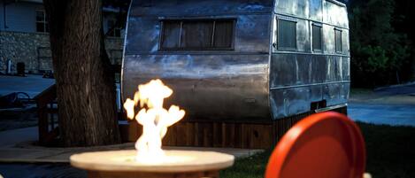 A vintage trailer with a fire in front of it, a tree, and a house in the background.