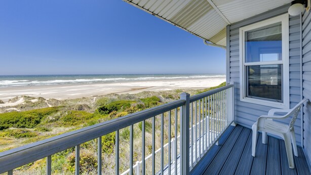 Eine Holzterrasse mit Geländer, ein weißer Plastikstuhl und Blick auf Strand und Dünen.