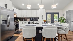A modern kitchen with white cabinets, a stainless steel refrigerator, a central island with bar stools, pendant lighting, and a view of the outdoors.