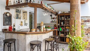 Un espace bar avec un comptoir en bois, des bouteilles et des tabourets. On y trouve des étagères remplies de divers objets et un panneau indiquant « BILLET ».
