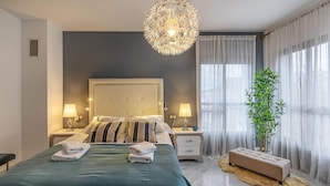 Une chambre à coucher moderne avec un grand lit, deux tables de chevet, un lustre décoratif, une plante en pot et des rideaux légers.
