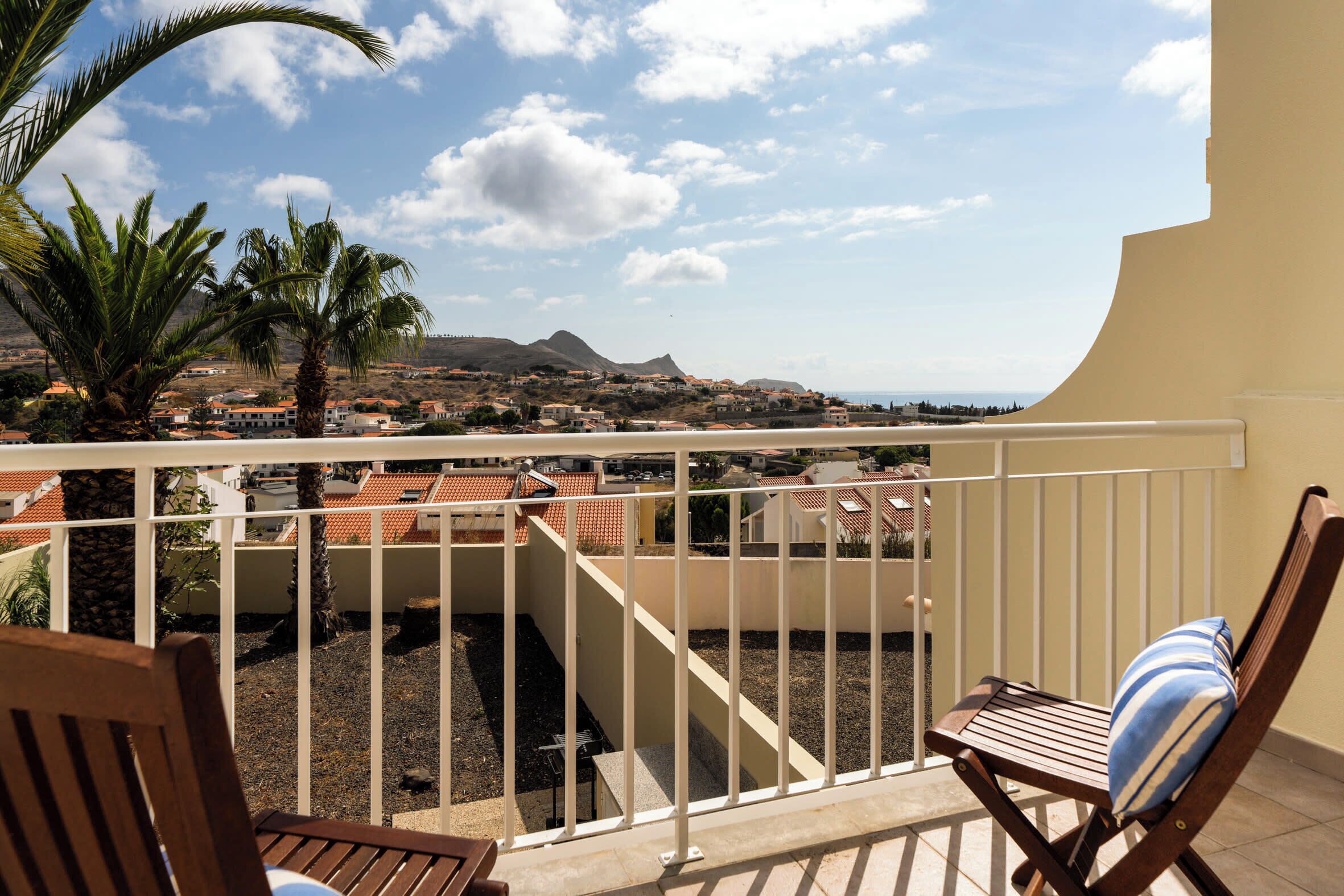 A balcony with a view of a coastal town, a palm tree, and a chair with a striped cushion.