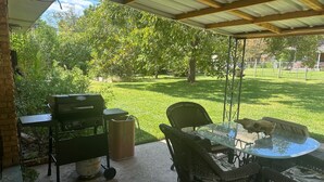 Een terras buiten met een barbecue, rieten meubels en een glazen tafel.