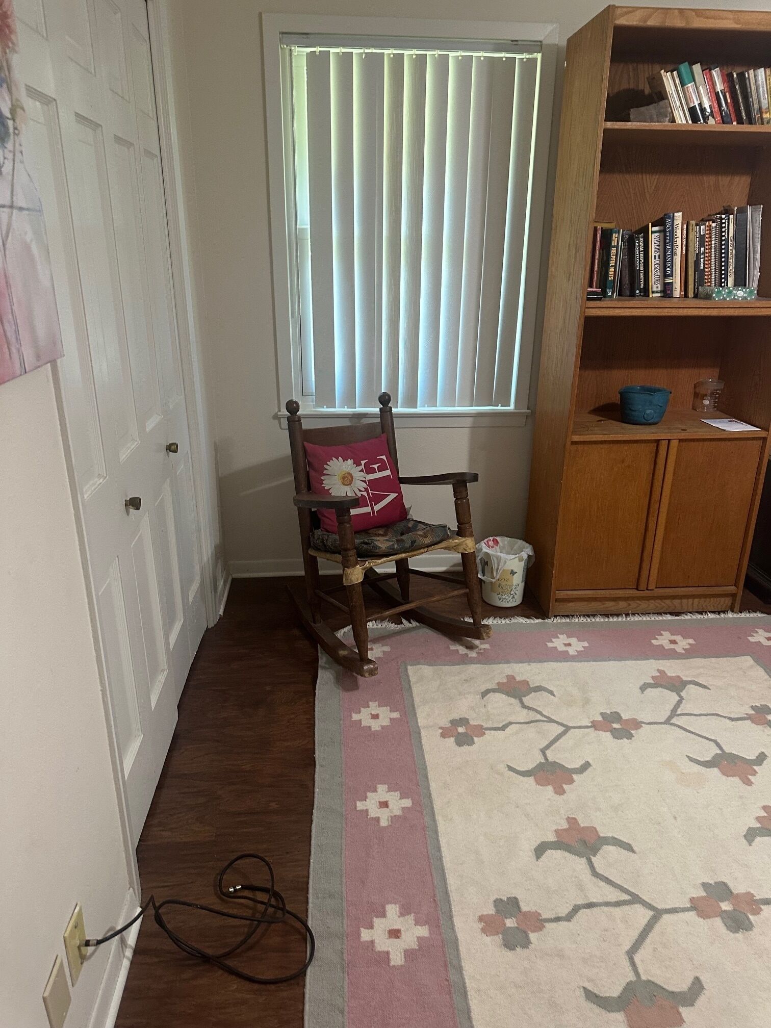 Ein Zimmer mit einem Holzstuhl, einem Bücherregal, einem Fenster mit Rollläden und einem gemusterten Teppich.