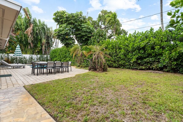 A patio area with a stone floor, outdoor furniture, and lush greenery including palm trees and bushes.