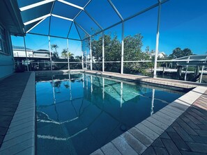 A clear indoor swimming pool with a glass ceiling, surrounded by a brick deck and a view of palm trees and buildings. - Spacious 4-Bed Retreat with Pool Short Stroll to the Beach (Fort Myers Beach)