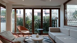 A modern living room with a sofa, a chair, a small table, and large windows offering a view of greenery and buildings. - Sense of Jedyod - near Maya Nimman (Chiang Mai)