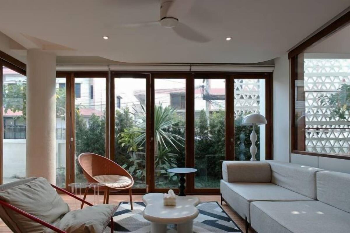 A modern living room with a sofa, a chair, a small table, and large windows offering a view of greenery and buildings.
