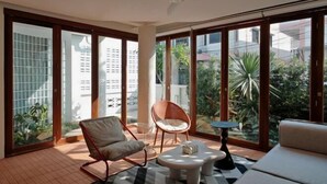 A modern living room with a sofa, two chairs, a coffee table, and a ceiling fan. Large windows offer views of greenery and buildings outside. - Sense of Jedyod - near Maya Nimman (Chiang Mai)