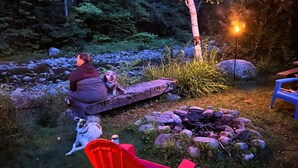 Eine Person sitzt auf einer Bank mit einem Hund, einem Feuerkorb und einem roten Stuhl in einem Garten.