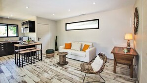 Ein modernes Wohnzimmer mit einer Küche, einem weißen Sofa mit orangefarbenen Kissen, einem Korbstuhl, einem kleinen Holztisch mit einer Lampe und einem großen Fenster, das für natürliches Licht sorgt.