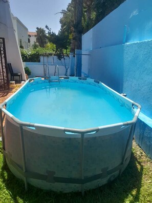 An outdoor swimming pool with a tiled deck, surrounded by a blue wall and a tree.