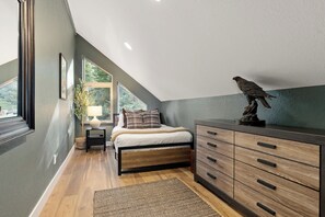 Een slaapkamer met een schuine plafond, een bed met houten frame, een dressoir met lades, een vloerkleed en een raam met uitzicht op groen.