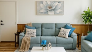 Un soggiorno con un divano grigio, due cuscini con la scritta "welcome HOME", un tavolino da caffè bianco, un vaso blu e un grande quadro floreale appeso al muro.