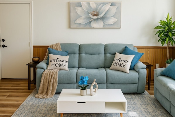 A living room with a grey sofa, two pillows with 'welcome HOME' text, a white coffee table, a blue vase, and a large floral painting on the wall.