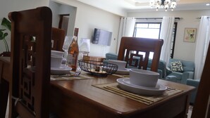 Un comedor con una mesa de madera dispuesta con platos, tazones y una botella. Se ve un televisor y un sofá al fondo.