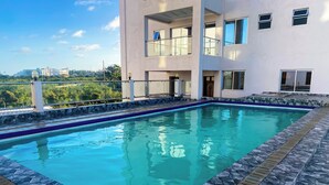 Una piscina rectangular con borde de piedra, junto a un edificio de varios pisos con balcones y ventanas amplias.