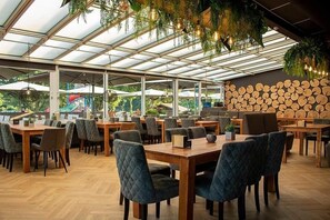 A spacious indoor dining area with wooden tables and chairs, a glass ceiling, and a wall of stacked logs. - Luxe chalet met ruime veranda & airco, vlak bij Toverland (Sevenum)