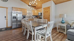 A modern kitchen with wooden cabinets, a stainless steel refrigerator, a dining table with chairs, and a hanging light fixture.