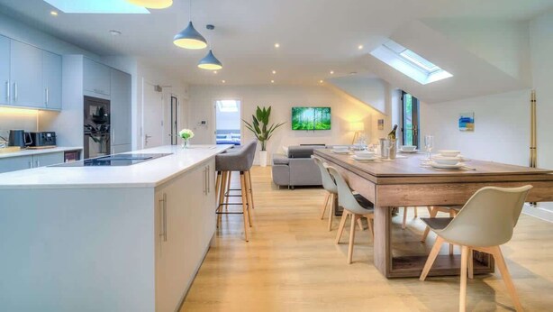 A modern kitchen with a central island, pendant lighting, and a dining area with a wooden table and chairs. - Penthouse Flat Manchester. 10 minutes to Airport. Sleeps 8. (Wythenshawe)