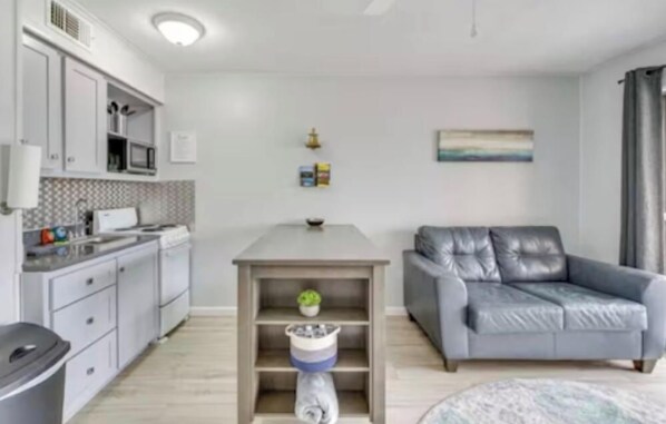 A modern kitchen with white cabinets, a microwave, and a sink. There's a grey sofa and a small table with a plant. - Beachside Escape (Corpus Christi)