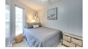 A bedroom with a bed, a bedside table, a lamp, a wall-mounted artwork, and a window. - Beachside Escape (Corpus Christi)