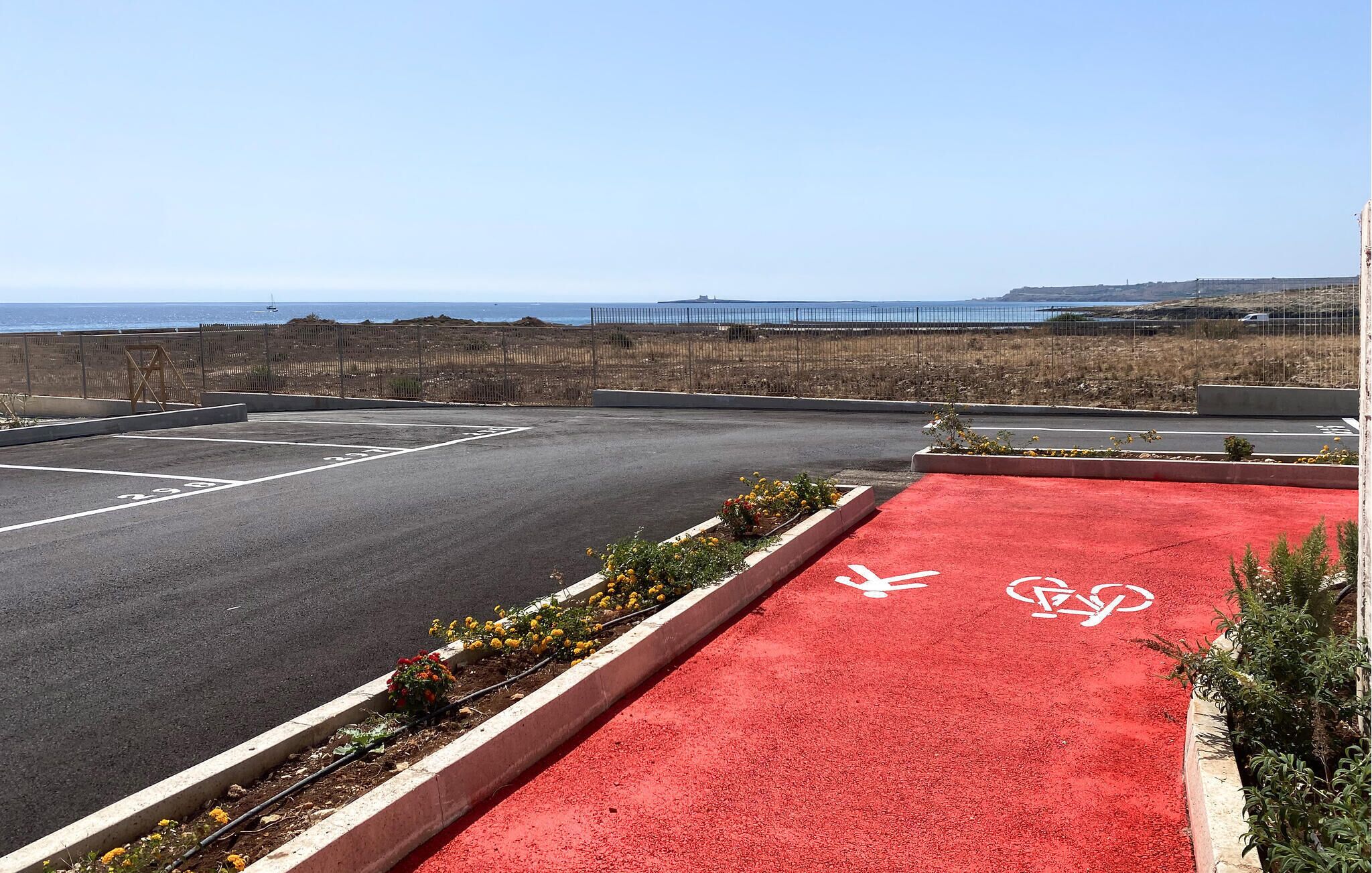 Ein Parkplatz mit markierten Stellplätzen, eine rote Fahrradspur und im Hintergrund Meer und Hügel.