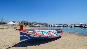 A beach with a rowboat on the sand, a clear blue sky, and a marina in the background. - Amazing apartment in Marzamemi with WiFi (Marzamemi)