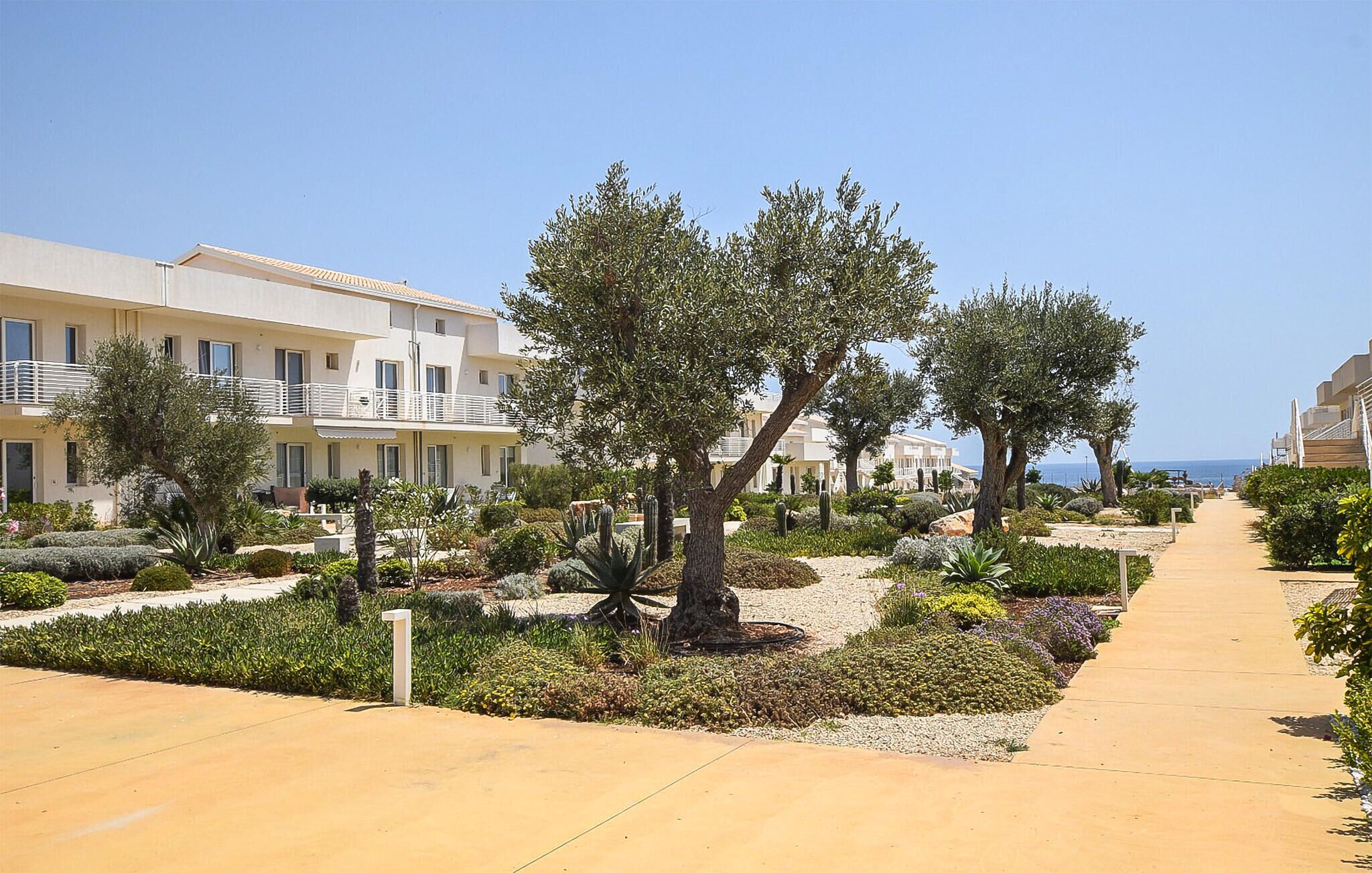 Un hotel frente al mar con un paseo empedrado, vegetación mediterránea y edificios con balcones con vistas al mar.