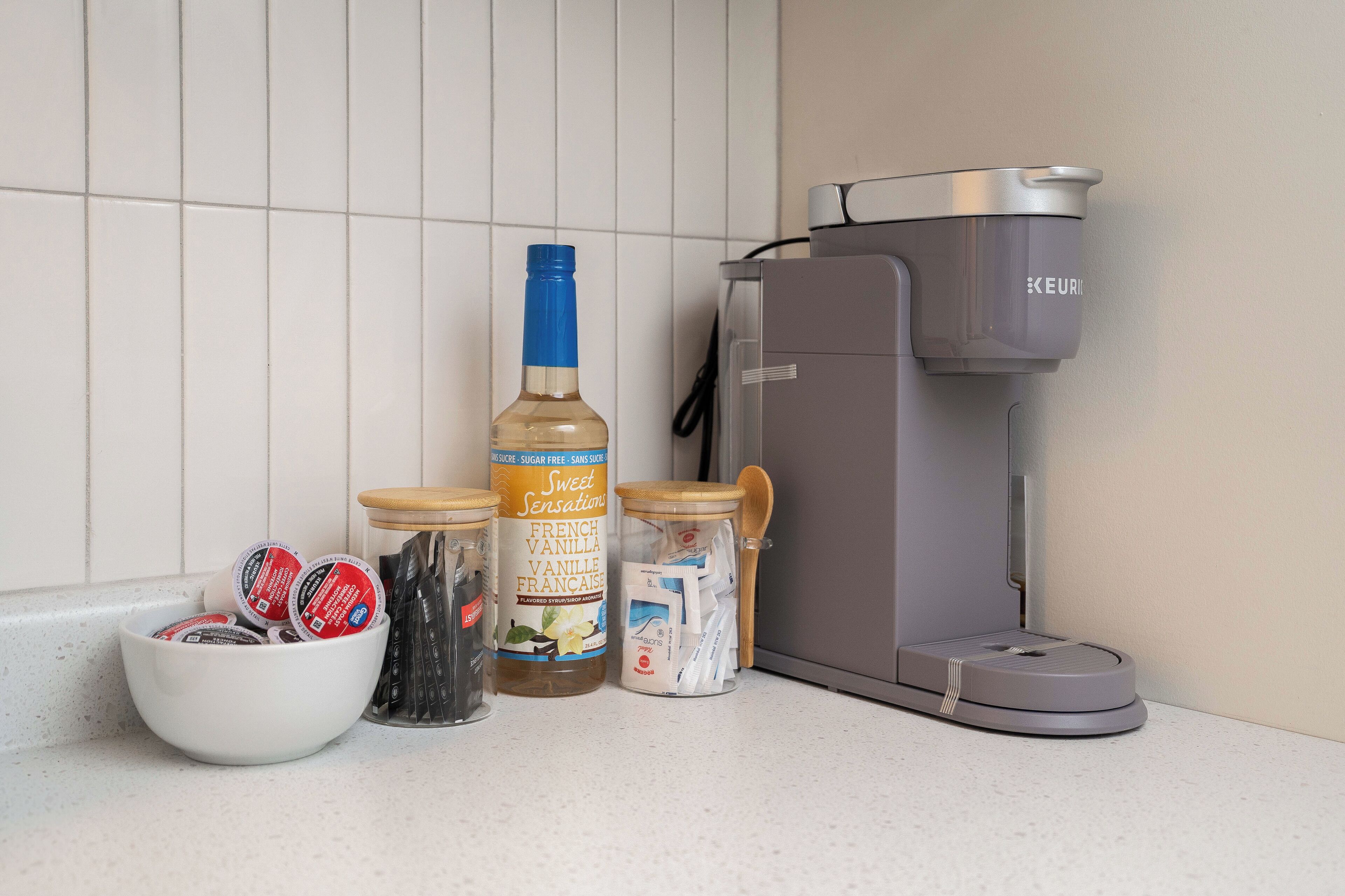 Una encimera de cocina con una cafetera, una botella de sirope de vainilla francés, y frascos de sobres de azúcar y crema para café.