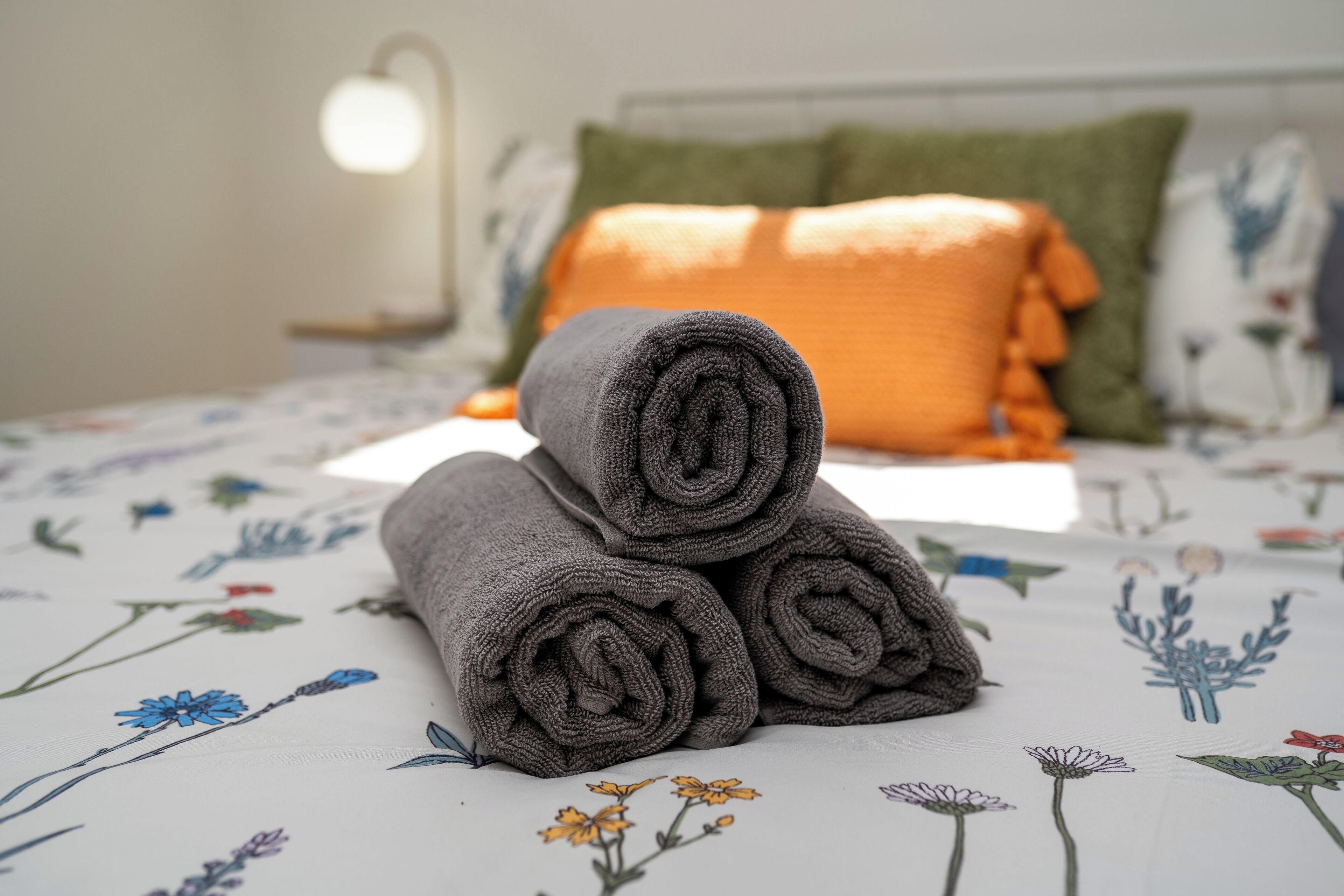 A neatly made bed with rolled towels, a green pillow, and a floral-patterned bedspread.